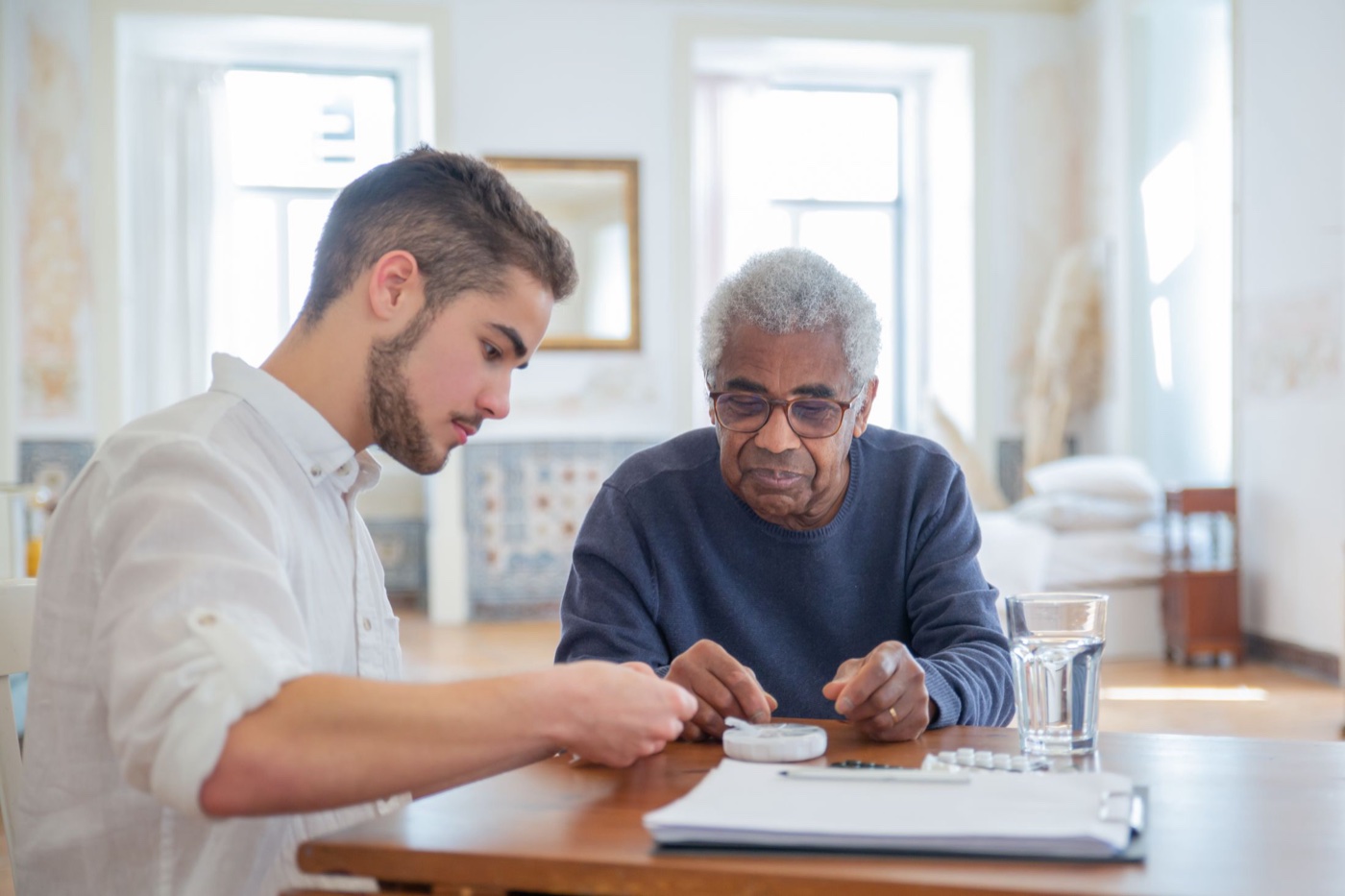 An adult grandson and his grandfather sort medications into a weekly organizer over the discharge paperwork at the kitchen table